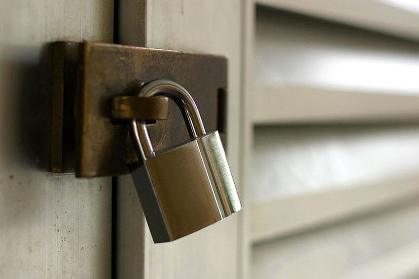 The image depicts a professional rubbish removal scene in a residential area of Bryncoch, with a focus on a light grey Hire Man and Van van positioned on a paved driveway. The van's side panel is visible, showing a smooth, reflective finish with minor dirt marks, suggesting recent use. In the immediate foreground, there is a large pile of mixed household waste, including black plastic rubbish bags, flattened cardboard boxes, and a few discarded wooden items, all stacked somewhat haphazardly on the concrete surface. To the right of the waste pile, a worker's hand, wearing a blue glove, is partially visible as they handle or prepare to load the debris into the van. The driveway is bordered by a low brick wall and some greenery, with a suburban residential building in the background, featuring a brick exterior and a tiled roof. Overcast daylight provides diffuse lighting, casting minimal shadows across the scene. The overall setting reflects a typical rubbish removal operation, highlighting the kind of service offered by Hire Man and Van in Bryncoch, CF32, with materials and context consistent with domestic clearance tasks within the local area.